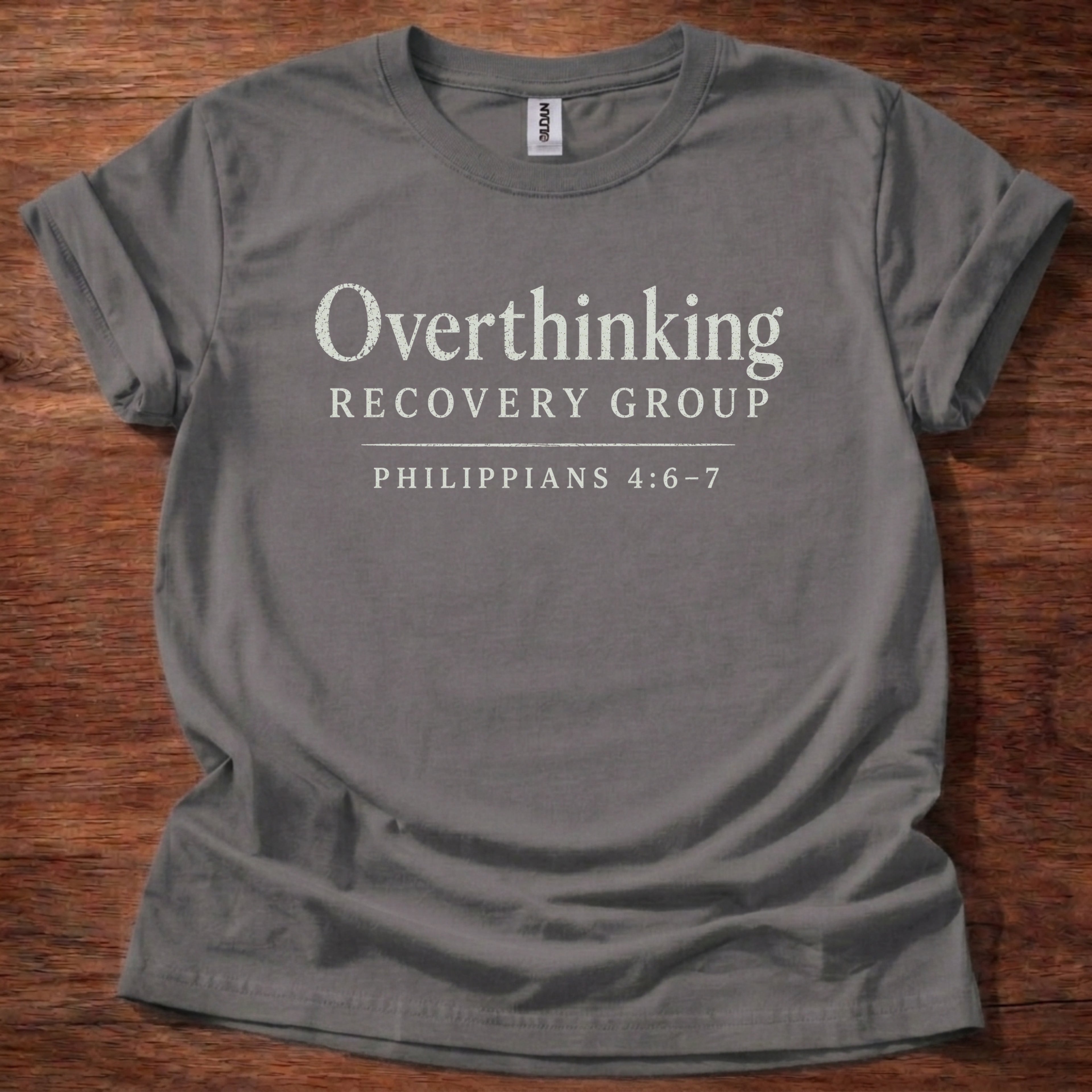 Gray t-shirt with 'Overthinking Recovery Group Philippians 4:6-7' text on a wooden surface