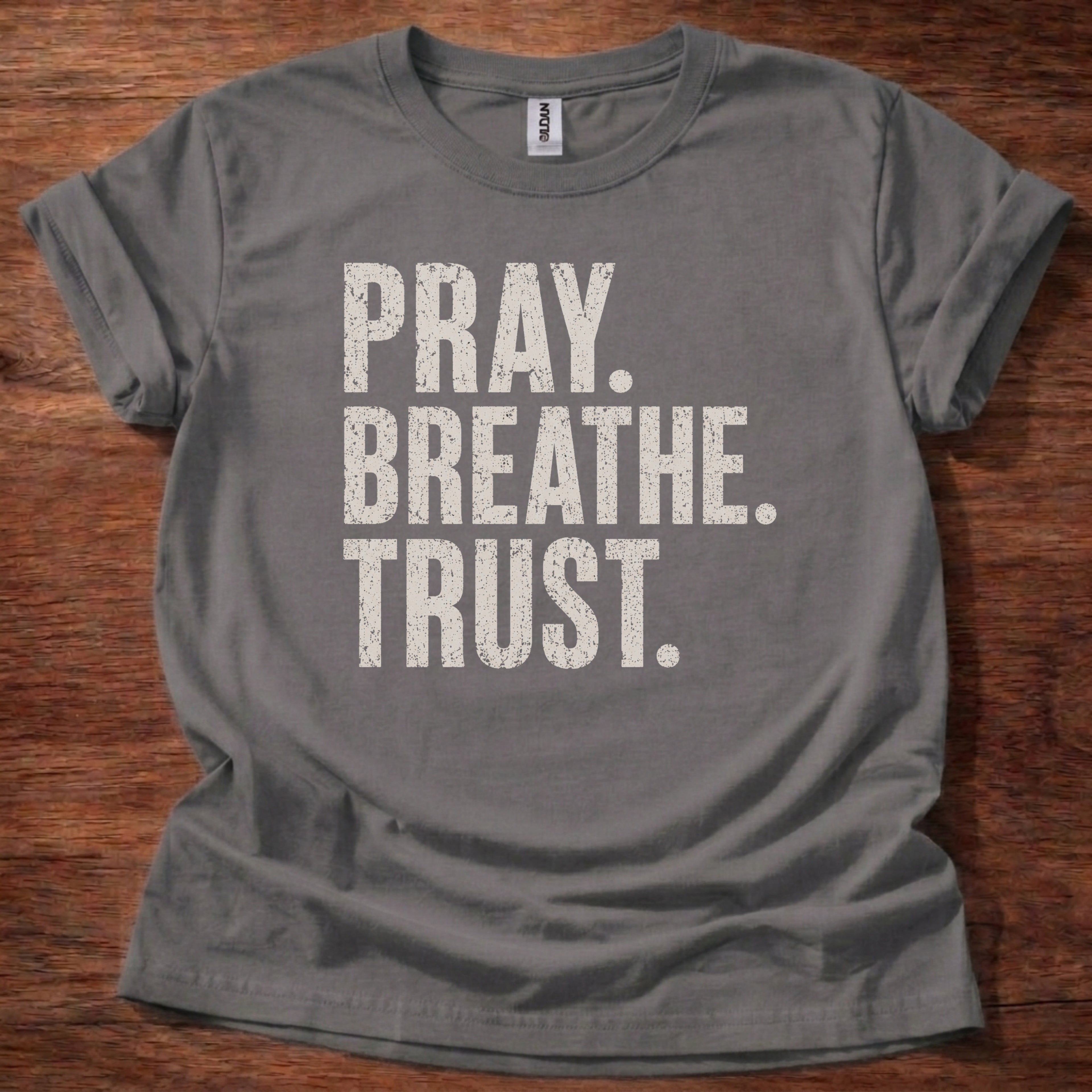 Gray t-shirt with 'PRAY. BREATHE. TRUST.' text on a wooden surface
