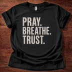 Black t-shirt with 'PRAY. BREATHE. TRUST.' text on a wooden surface