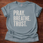 Blue t-shirt with 'PRAY. BREATHE. TRUST.' text on a wooden surface