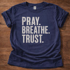 Navy blue t-shirt with 'PRAY. BREATHE. TRUST.' text on a wooden surface