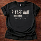 Black t-shirt with 'PLEASE WAIT. PSALM 46:10' text on a wooden surface