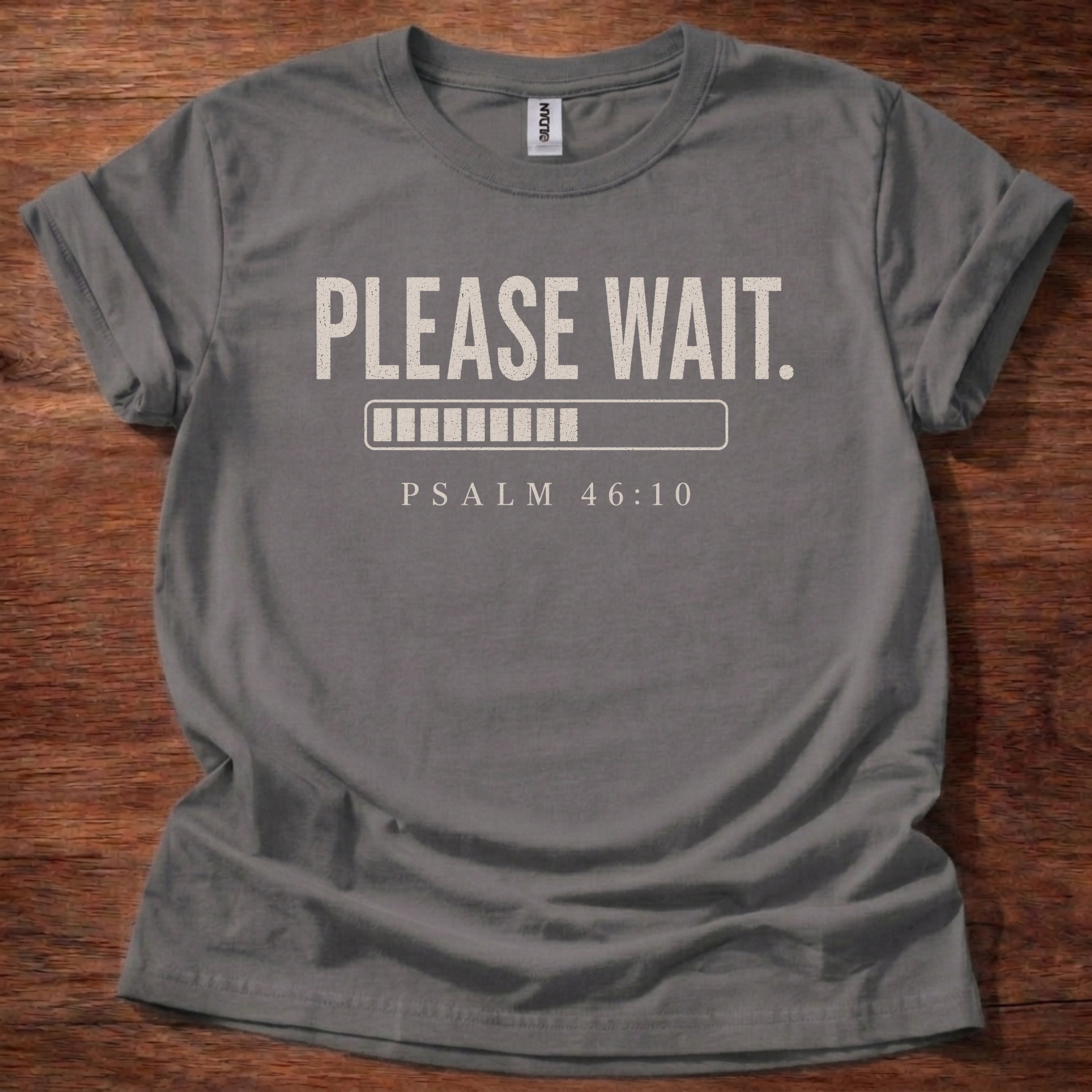 Gray t-shirt with 'PLEASE WAIT. PSALM 46:10' text on a wooden surface