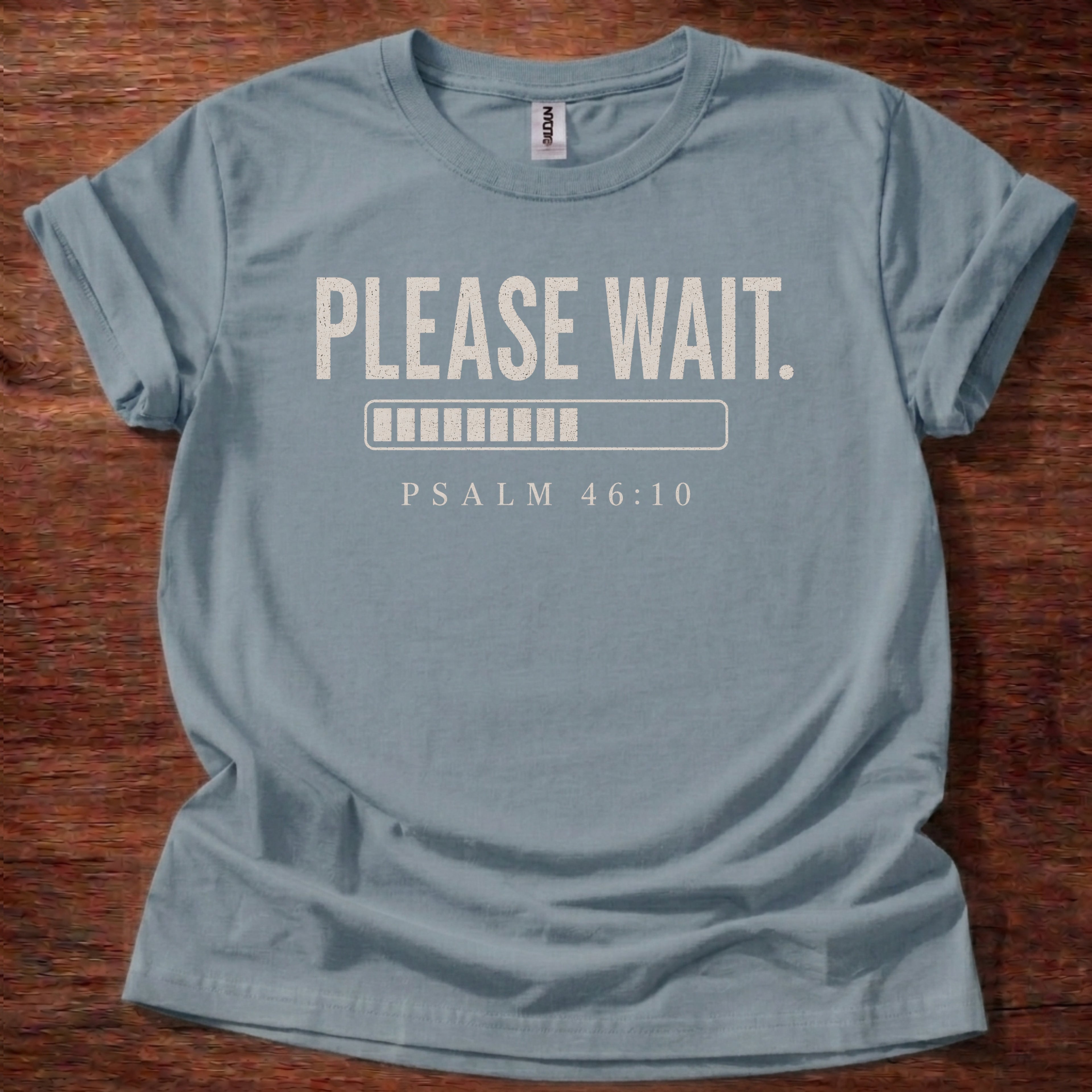 Blue t-shirt with 'Please Wait' and Psalm 46:10 text on a wooden surface