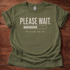 Green t-shirt with 'PLEASE WAIT. PSALM 46:10' text on a wooden surface