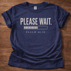 Navy blue t-shirt with 'PLEASE WAIT. PSALM 46:10' text on a wooden surface