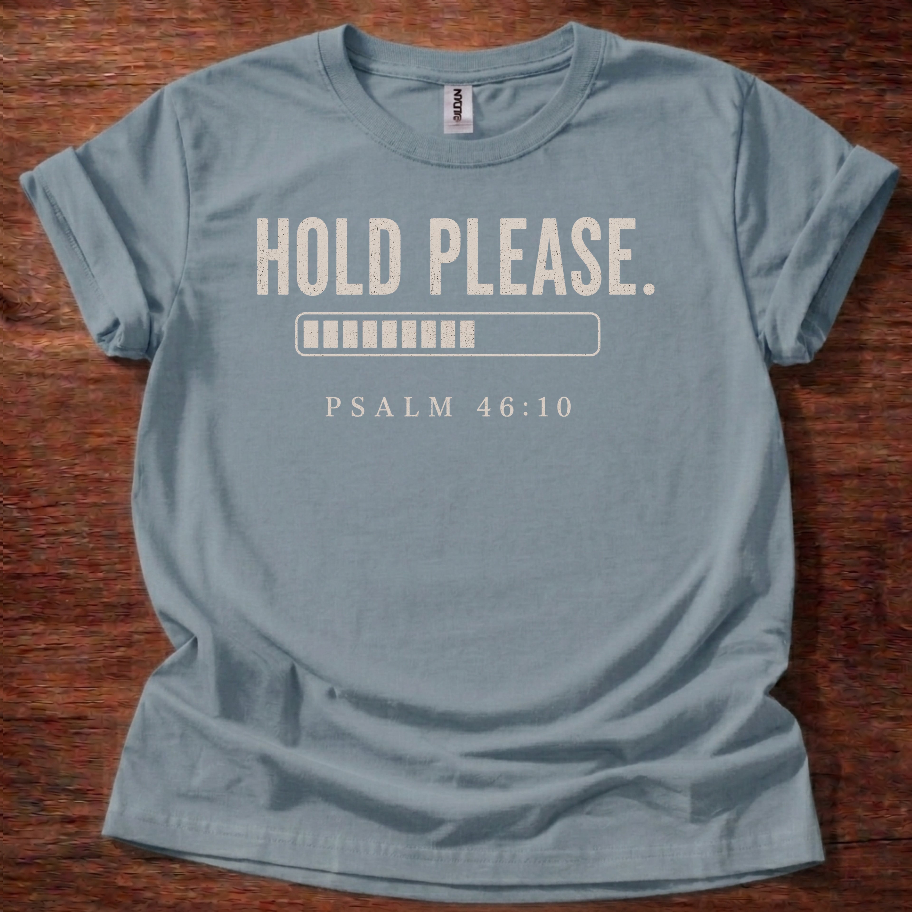 Blue t-shirt with 'Hold Please. Psalm 46:10' text on a wooden surface