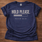 Navy blue t-shirt with 'Hold Please. Psalm 46:10' text on a wooden surface