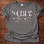 Gray t-shirt with 'Your Mind Needs Rest Too' text on a wooden surface