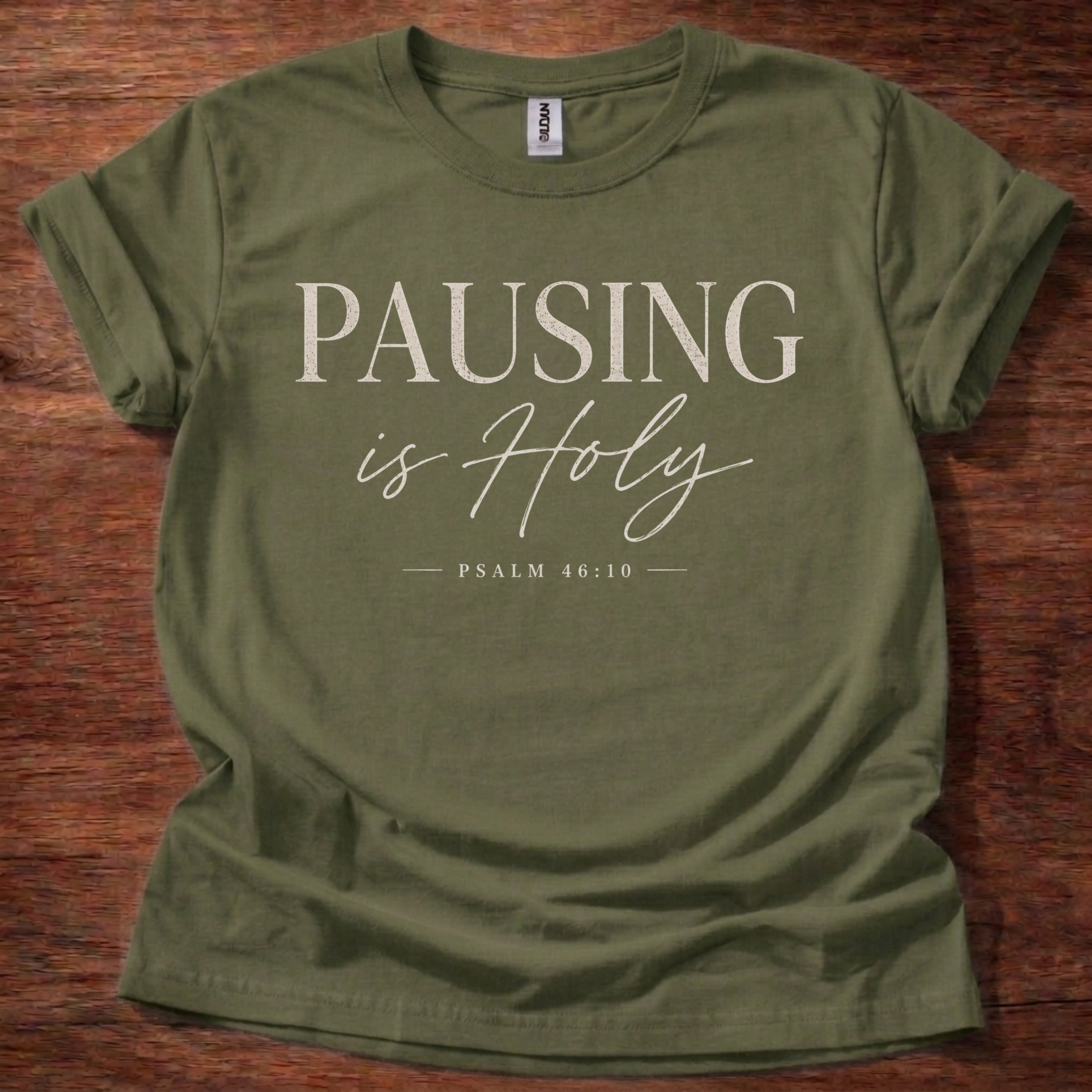 Green t-shirt with 'PAUSING is Holy' text on a wooden surface