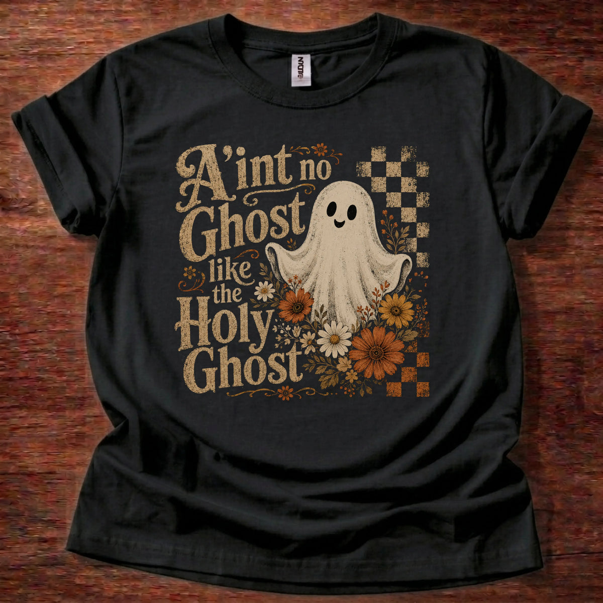 A black short-sleeve graphic t-shirt laid flat on a dark wood background. The shirt features a vintage-style Halloween graphic with the text 'A'int no Ghost like the Holy Ghost' in a distressed beige font, a white cartoon ghost, orange and yellow flowers, and a checkered pattern.