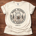 An off-white short-sleeve t-shirt laid flat on a dark wood background, featuring a circular graphic of a raccoon in a plaid sweater holding a mug, surrounded by the text 'BASIC. BLESSED BRING THE PUMPKIN SPICE' and 'JAMES 1:17' below.
