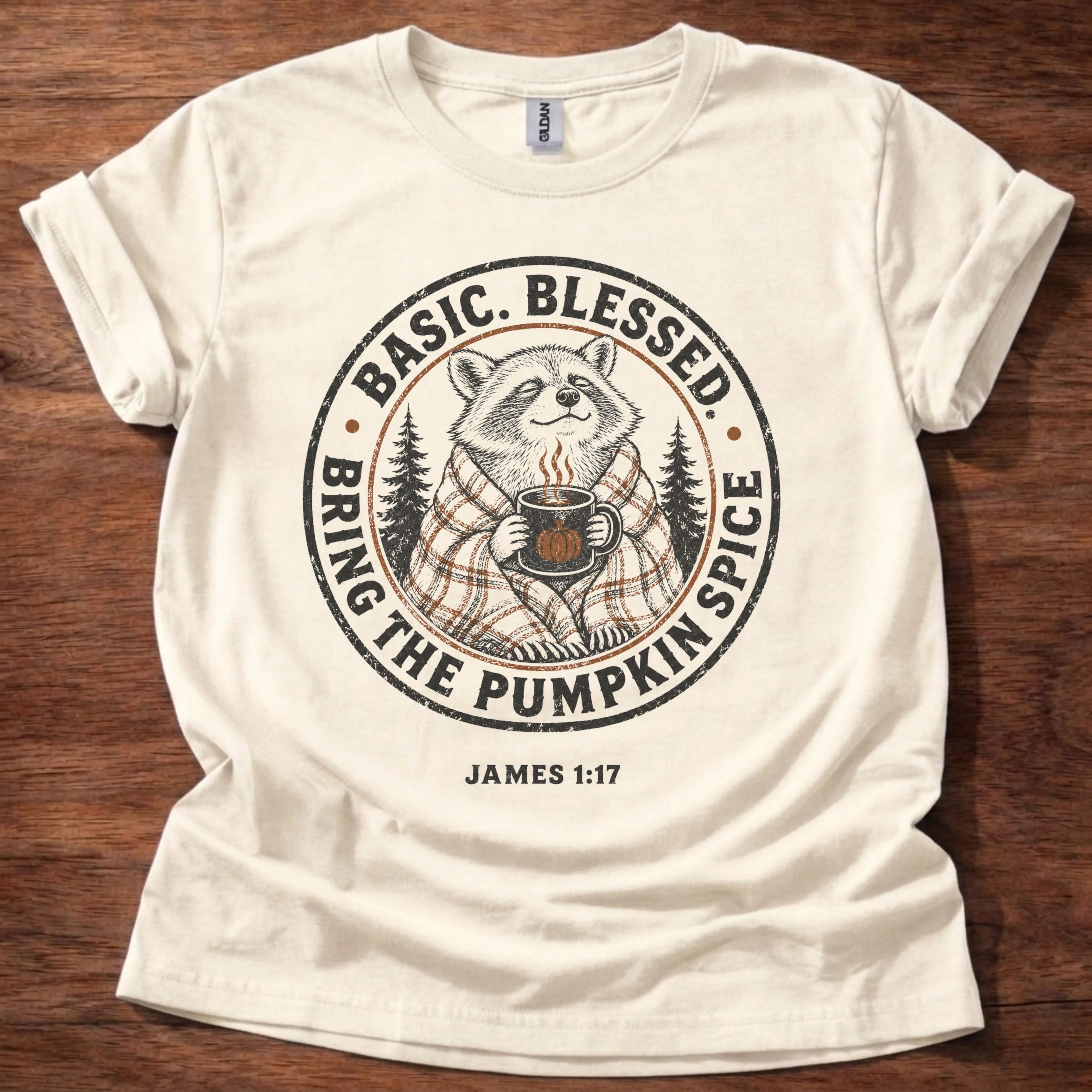 An off-white short-sleeve t-shirt laid flat on a dark wood background, featuring a circular graphic of a raccoon in a plaid sweater holding a mug, surrounded by the text 'BASIC. BLESSED BRING THE PUMPKIN SPICE' and 'JAMES 1:17' below.
