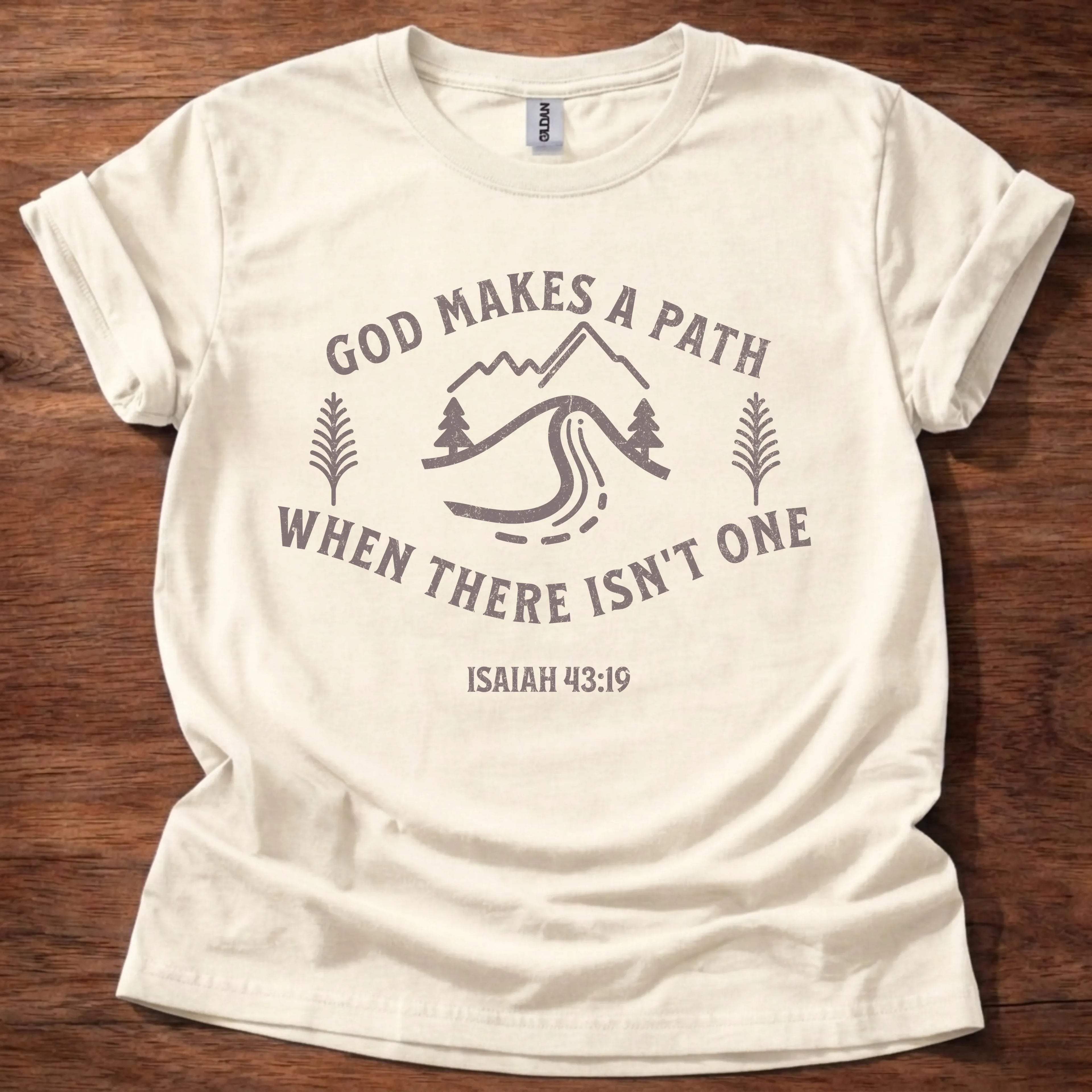 Mauve mountain, river, and pine tree illustration t-shirt with the text 'God Makes A Path When There Isn't One' and Isaiah 43:19