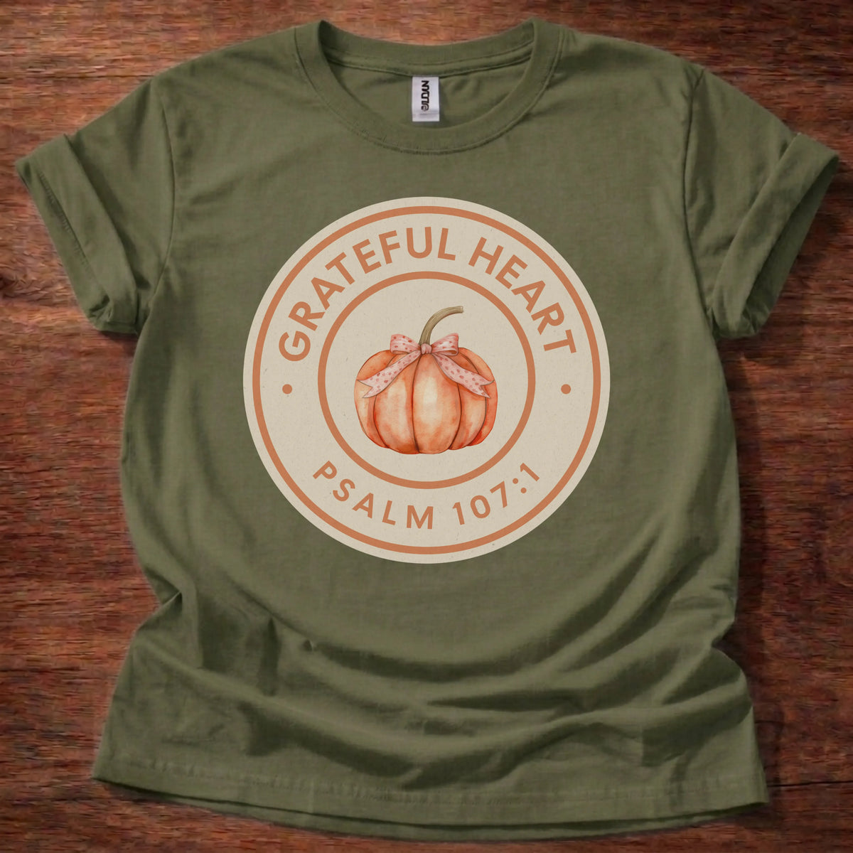 Circular badge t-shirt with a watercolor pumpkin illustration and the text 'Grateful Heart' and 'Psalm 107:1' arched around the ring