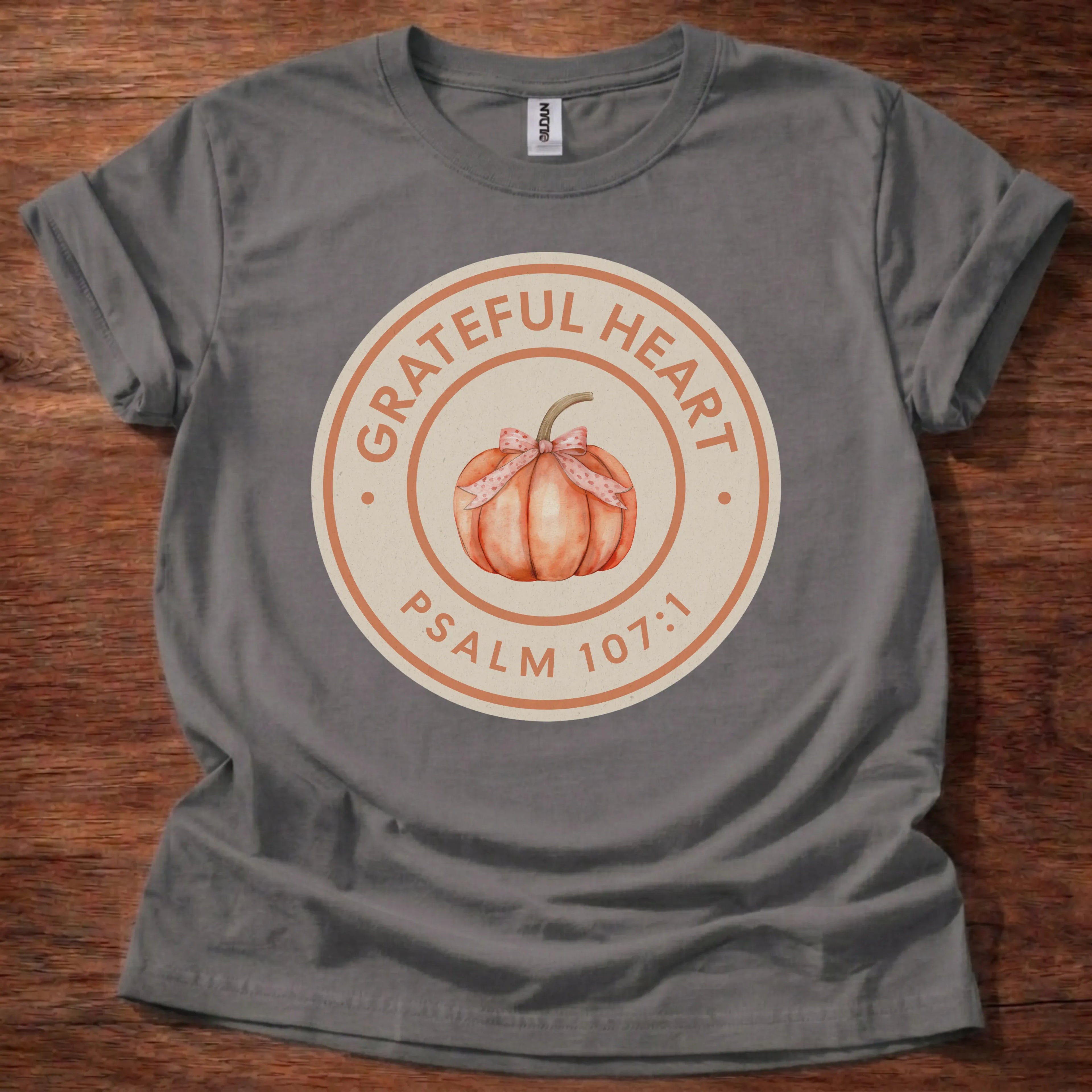 Circular badge t-shirt with a watercolor pumpkin illustration and the text 'Grateful Heart' and 'Psalm 107:1' arched around the ring