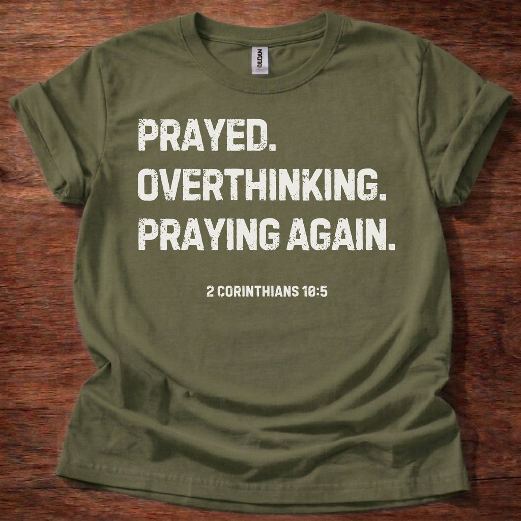 Green t-shirt with white text that reads Prayed. Overthinking. Praying again on a wooden surface