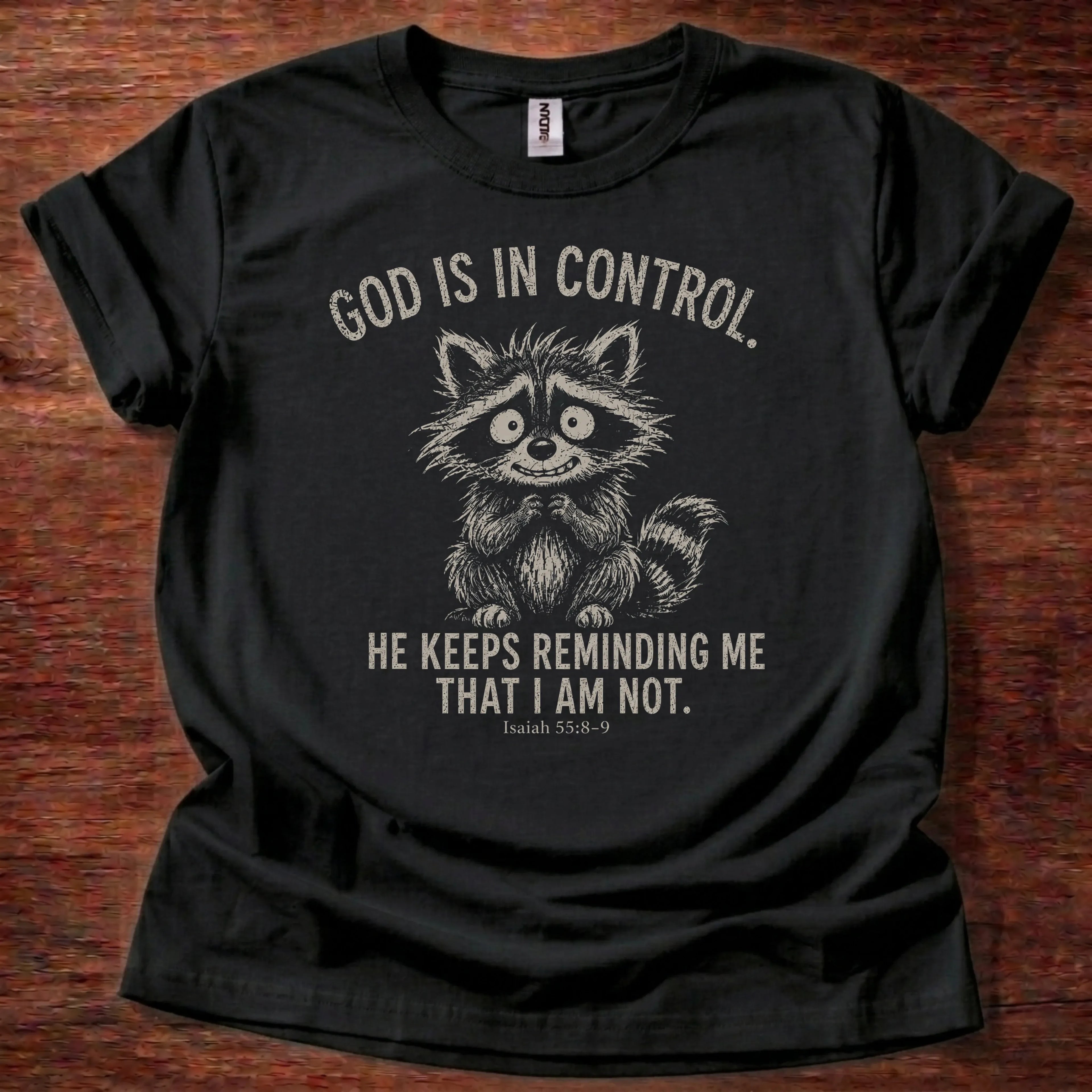 White fine-line raccoon illustration t-shirt with the text 'God Is In Control. He Keeps Reminding Me That I Am Not.' and Isaiah 55:8-9