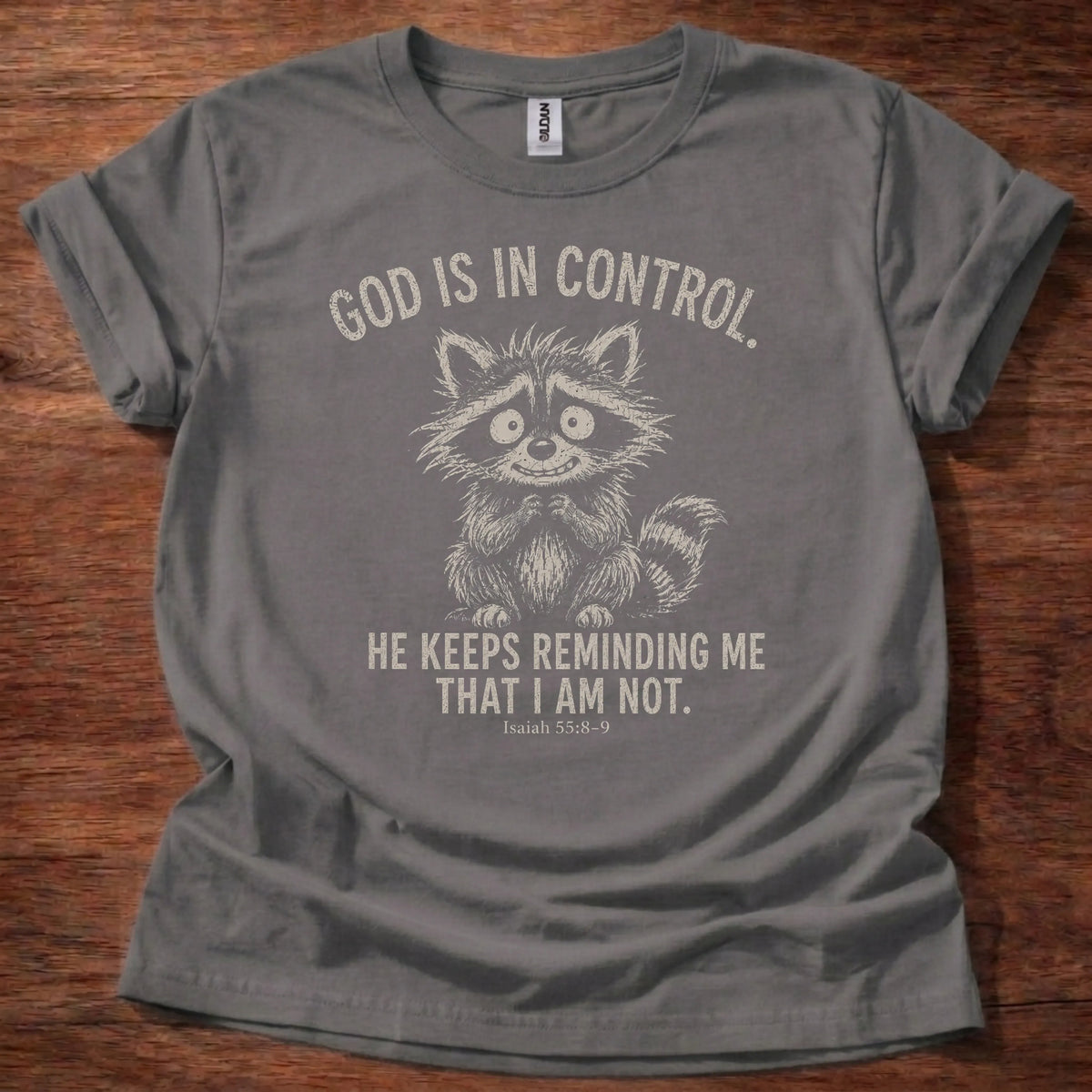 White fine-line raccoon illustration t-shirt with the text 'God Is In Control. He Keeps Reminding Me That I Am Not.' and Isaiah 55:8-9