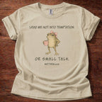 A beige graphic t-shirt laid flat on a dark brown wooden surface. The shirt features a printed illustration of a cartoon frog wearing a pink bow and holding a pink mug, with the text 'LEAD ME NOT INTO TEMPTATION OR SMALL TALK MATTHEW 6:13'.