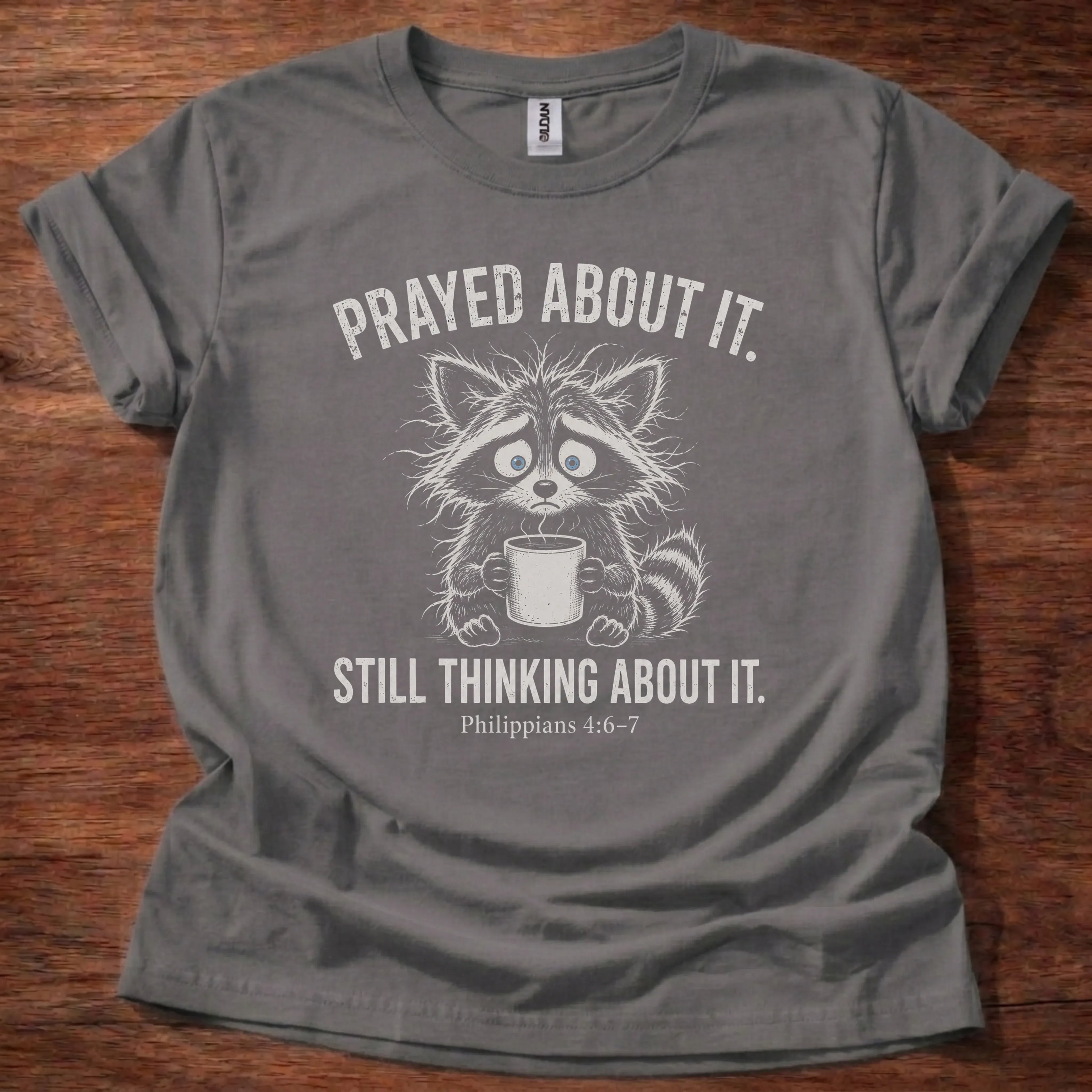 White fine-line raccoon illustration holding a mug t-shirt with the text 'Prayed About It. Still Thinking About It.' and Philippians 4:6-7