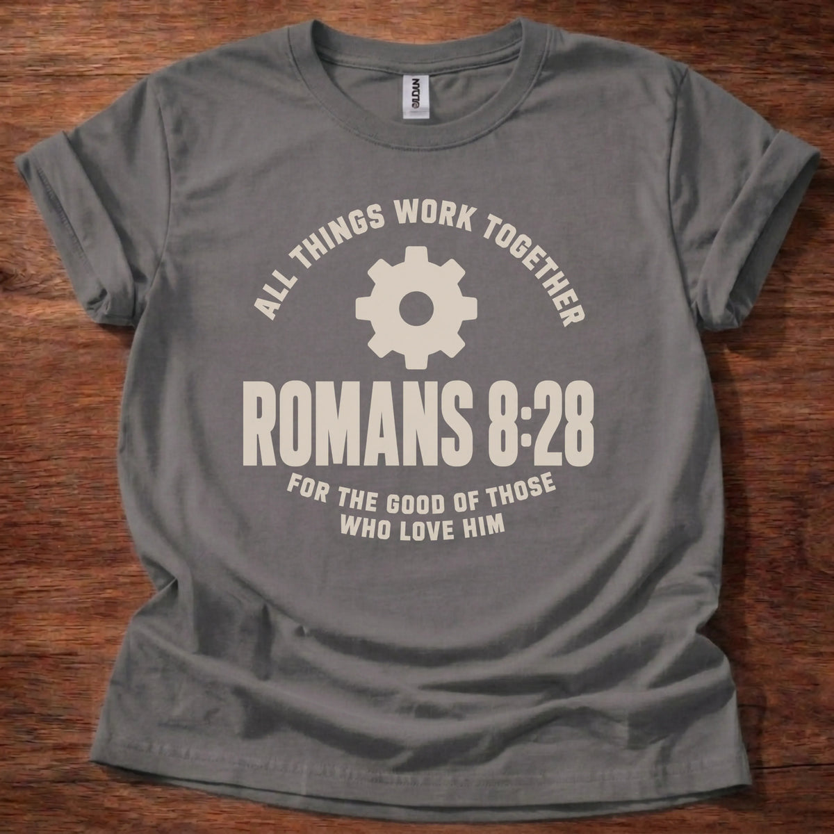 Cream gear icon t-shirt with the text 'All Things Work Together' arched above and 'Romans 8:28' below