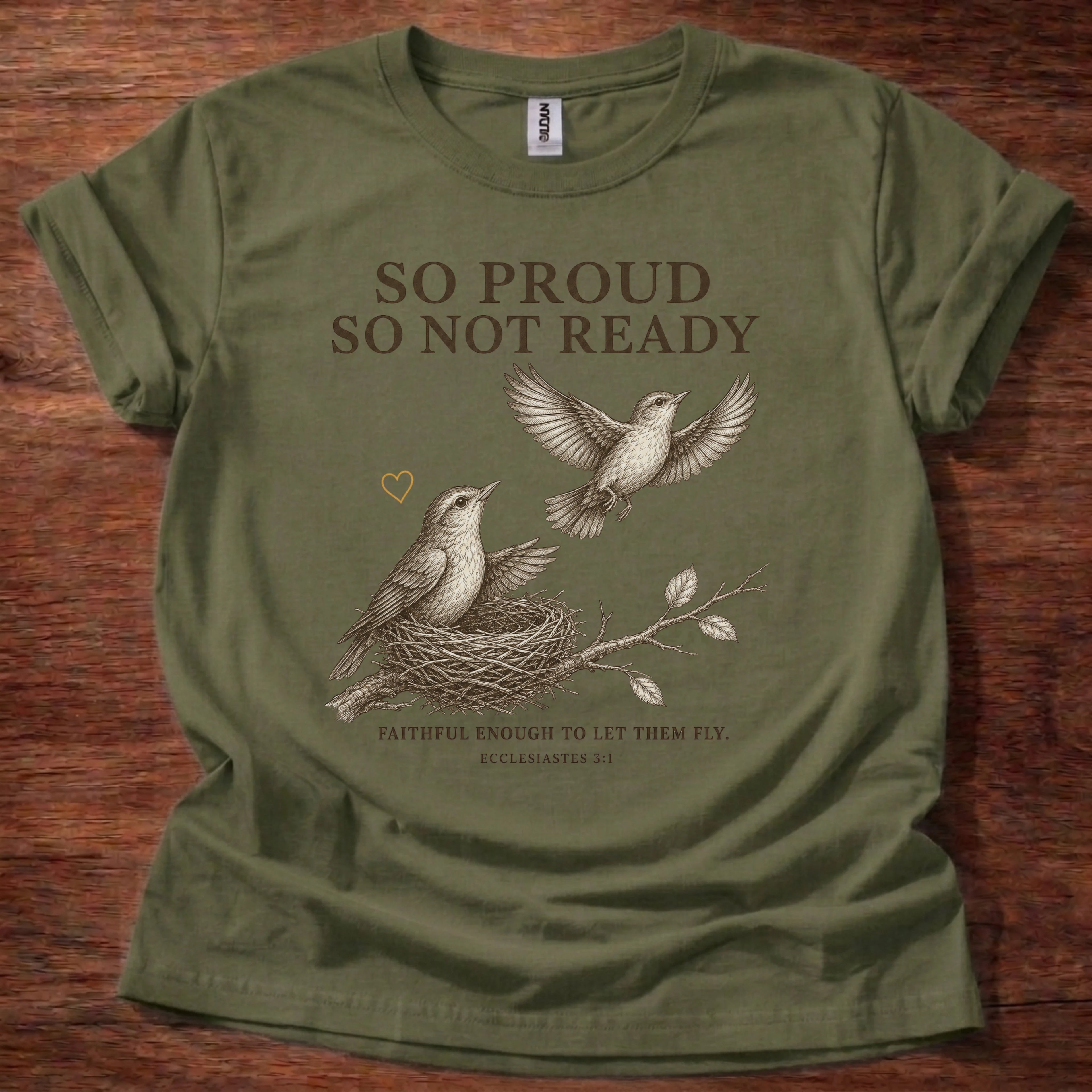 An olive green short-sleeve t-shirt with a graphic print of two birds and a nest, featuring the text 'SO PROUD SO NOT READY' and 'FAITHFUL ENOUGH TO LET THEM FLY.' laid flat on a dark wood background.