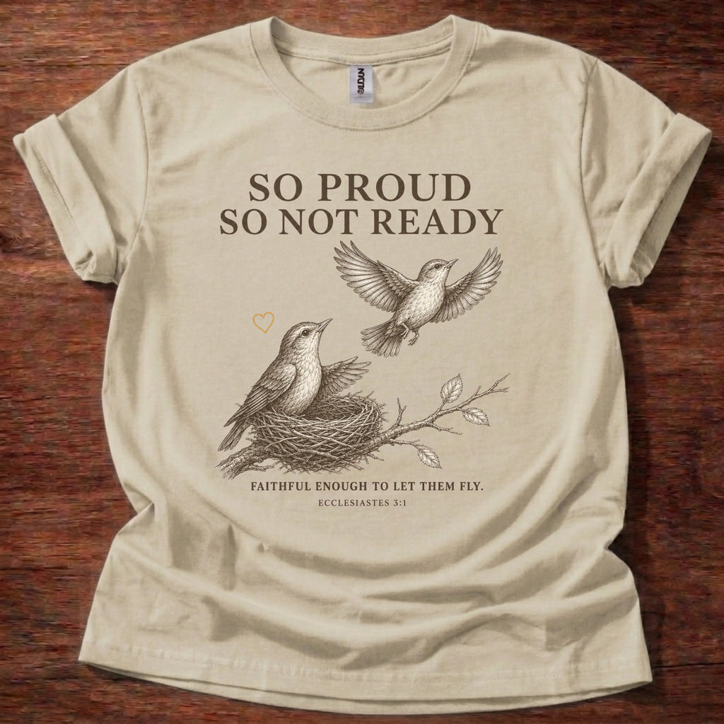 A beige short-sleeve graphic T-shirt laid flat on a dark wood background. The shirt features a printed design of two birds and a nest with the text 'SO PROUD SO NOT READY' and 'FAITHFUL ENOUGH TO LET THEM FLY.
