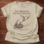 A beige short-sleeve graphic T-shirt laid flat on a dark wood background. The shirt features a printed design of two birds and a nest with the text 'SO PROUD SO NOT READY' and 'FAITHFUL ENOUGH TO LET THEM FLY.