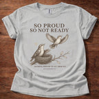 A light gray short-sleeve t-shirt with a brown graphic print of two birds and a nest, featuring the text 'SO PROUD SO NOT READY' and 'FAITHFUL ENOUGH TO LET THEM FLY. ECCLESIASTES 3:1', laid flat on a dark wood background.