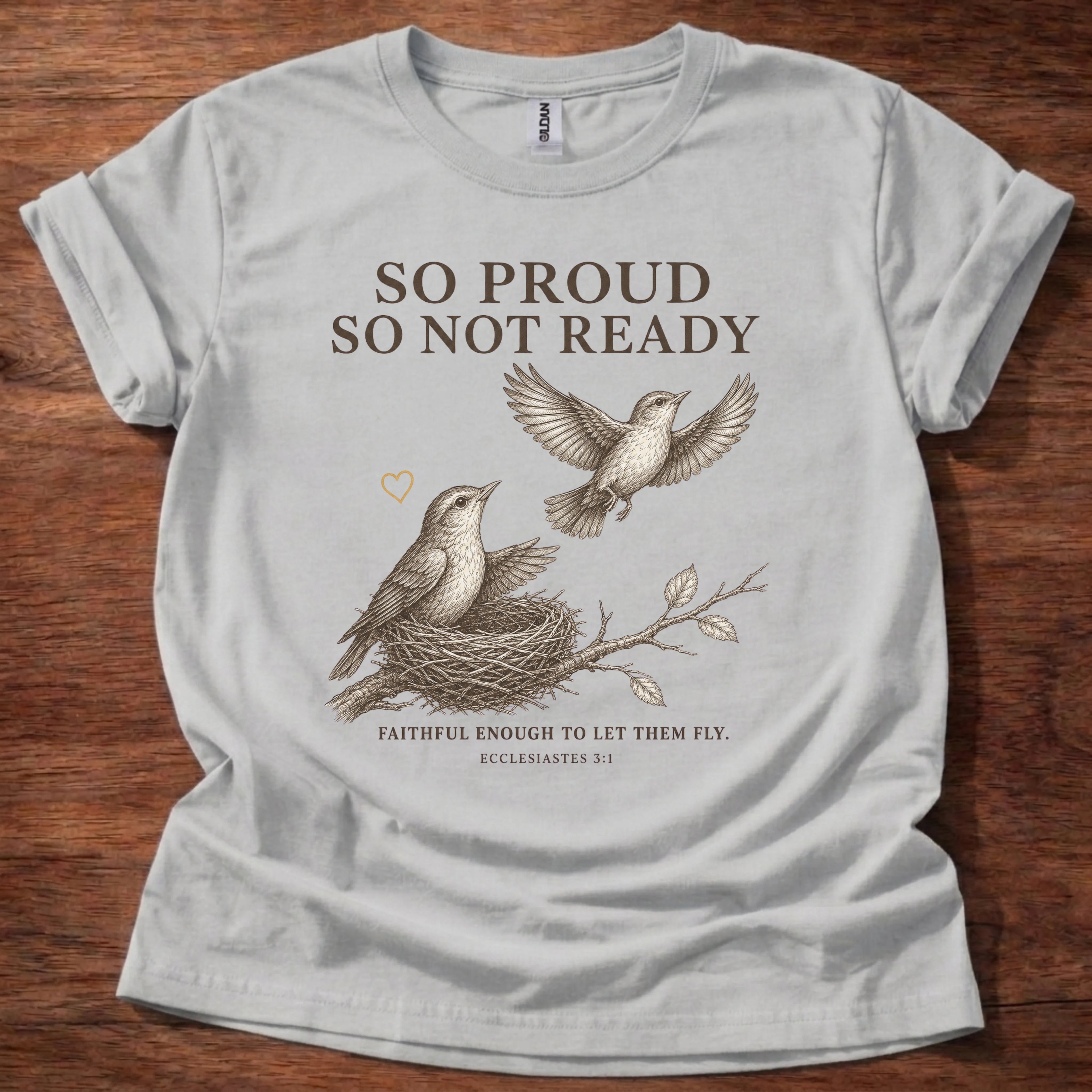 A light gray short-sleeve t-shirt with a brown graphic print of two birds and a nest, featuring the text 'SO PROUD SO NOT READY' and 'FAITHFUL ENOUGH TO LET THEM FLY. ECCLESIASTES 3:1', laid flat on a dark wood background.