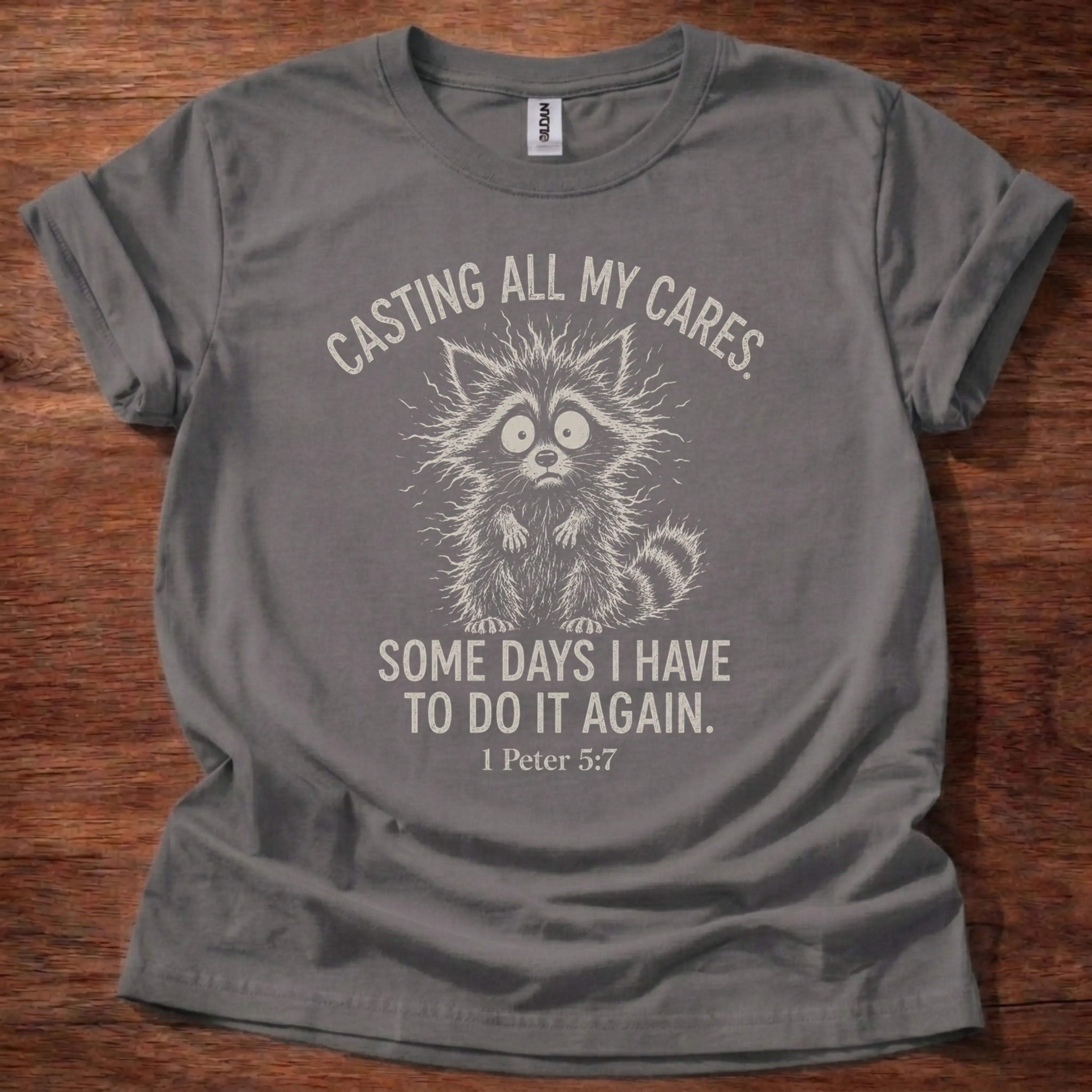 White fine-line raccoon illustration t-shirt with the text 'Casting All My Cares. Some Days I Have To Do It Again.' and 1 Peter 5:7