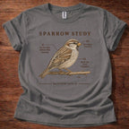 A gray short-sleeve t-shirt with a brown and beige printed graphic of a sparrow perched on a branch, with the text 'SPARROW STUDY' and scripture reference 'MATTHEW 10:29-31'. The shirt is laid flat on a dark wood background.