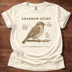 An off-white short-sleeve t-shirt laid flat on a dark wood background, featuring a vintage-style sparrow illustration with the text 'SPARROW STUDY' and biblical reference 'MATTHEW 10:29-31'.