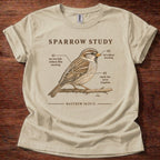 A beige short-sleeve crewneck t-shirt featuring a vintage-style brown graphic of a sparrow perched on a branch with text labels and the Bible verse Matthew 10:29-31, displayed flat on a dark wood background.