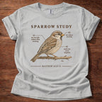 A light gray graphic t-shirt laid flat on a dark wood background. The shirt features a detailed illustration of a brown sparrow perched on a branch, with humorous text labels pointing to its features, and the quote 'MATTHEW 10:29-31' printed below.