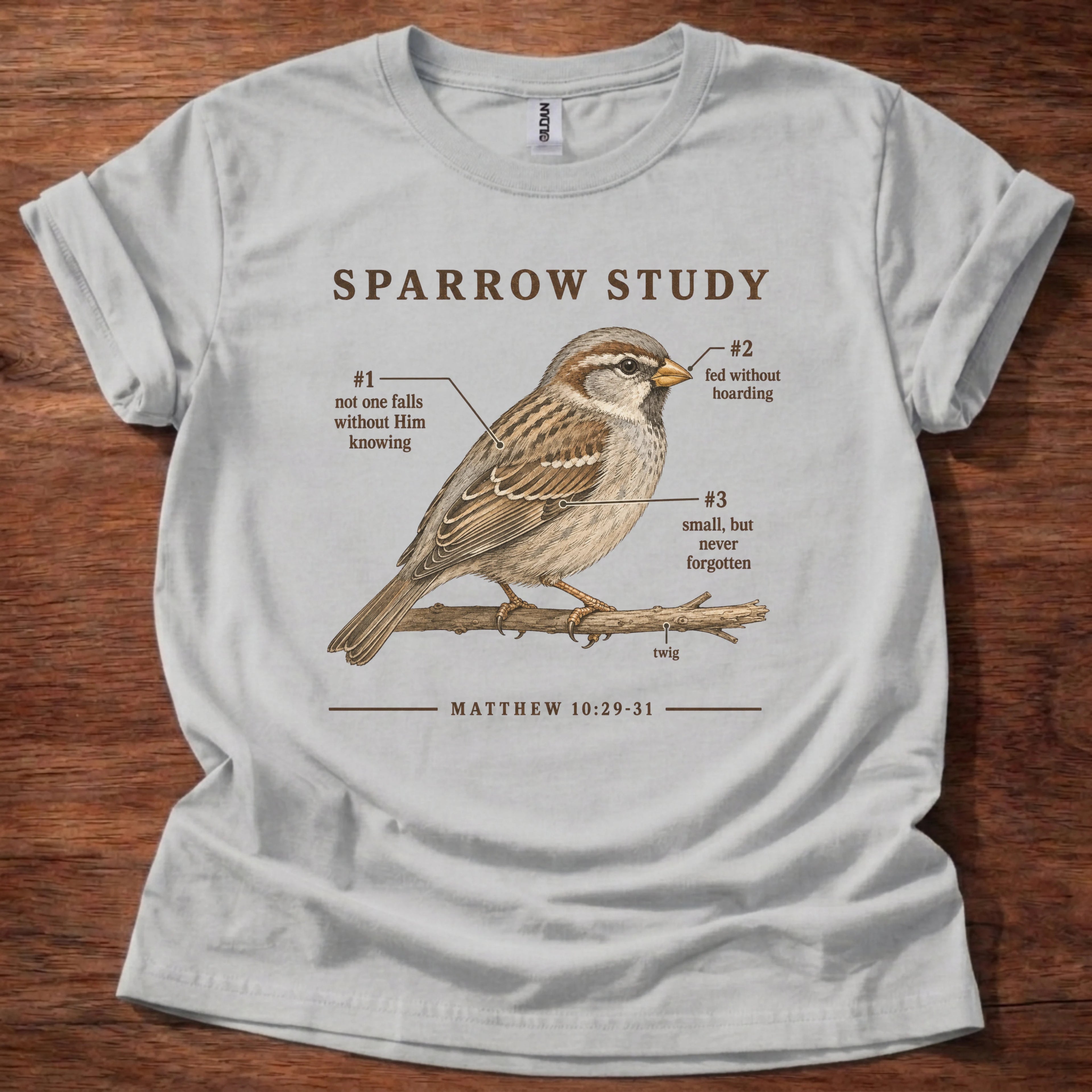 A light gray graphic t-shirt laid flat on a dark wood background. The shirt features a detailed illustration of a brown sparrow perched on a branch, with humorous text labels pointing to its features, and the quote 'MATTHEW 10:29-31' printed below.