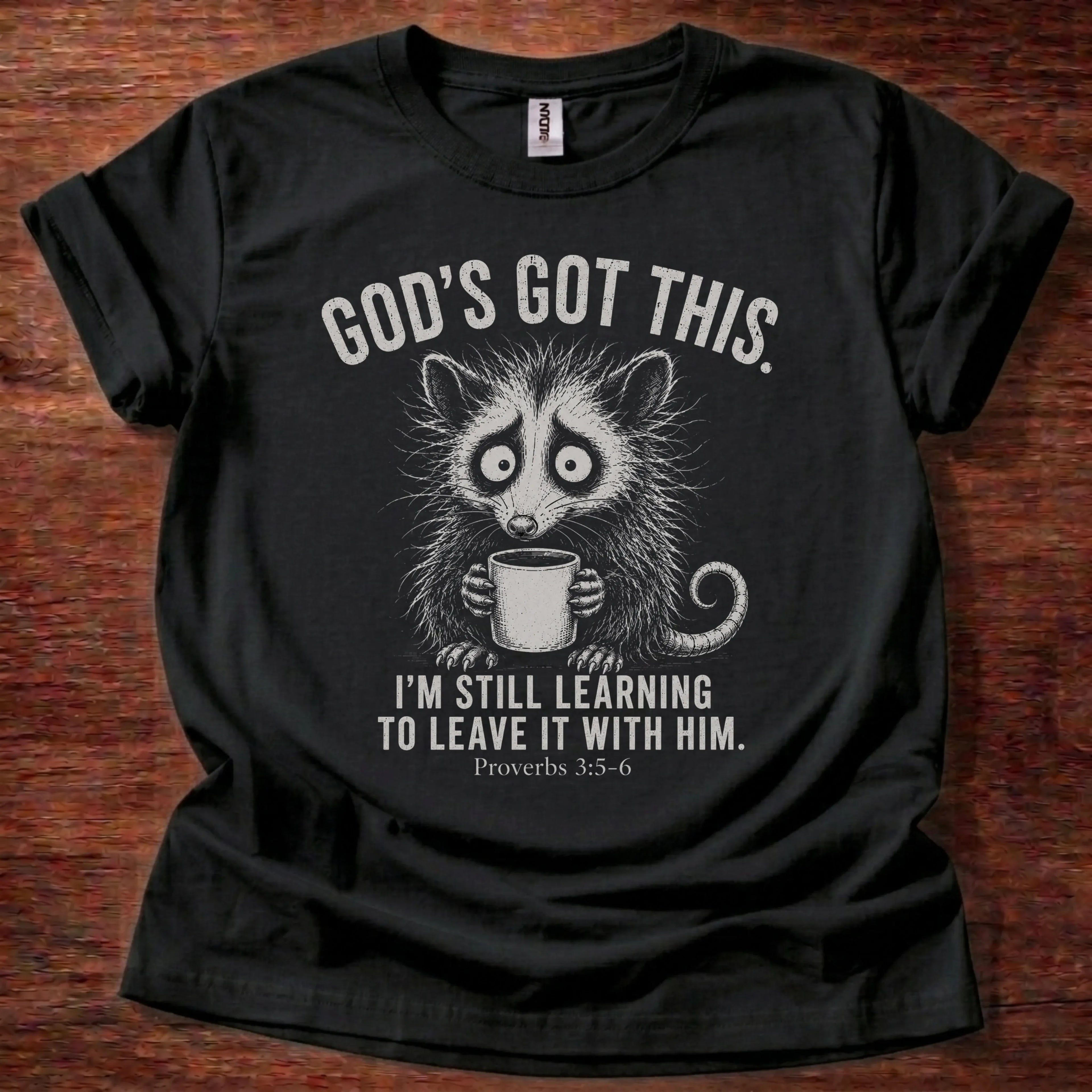 White fine-line raccoon illustration holding a coffee mug t-shirt with the text 'God's Got This. I'm Still Learning To Leave It With Him.' and Proverbs 3:5-6