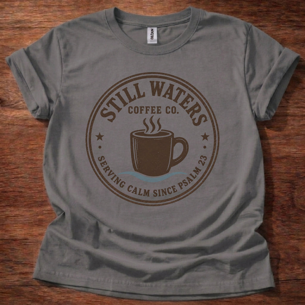 A gray short-sleeve crewneck t-shirt laid flat on a dark wood background, featuring a circular brown graphic print that reads 'Still Waters Coffee Co.' with a coffee cup illustration.