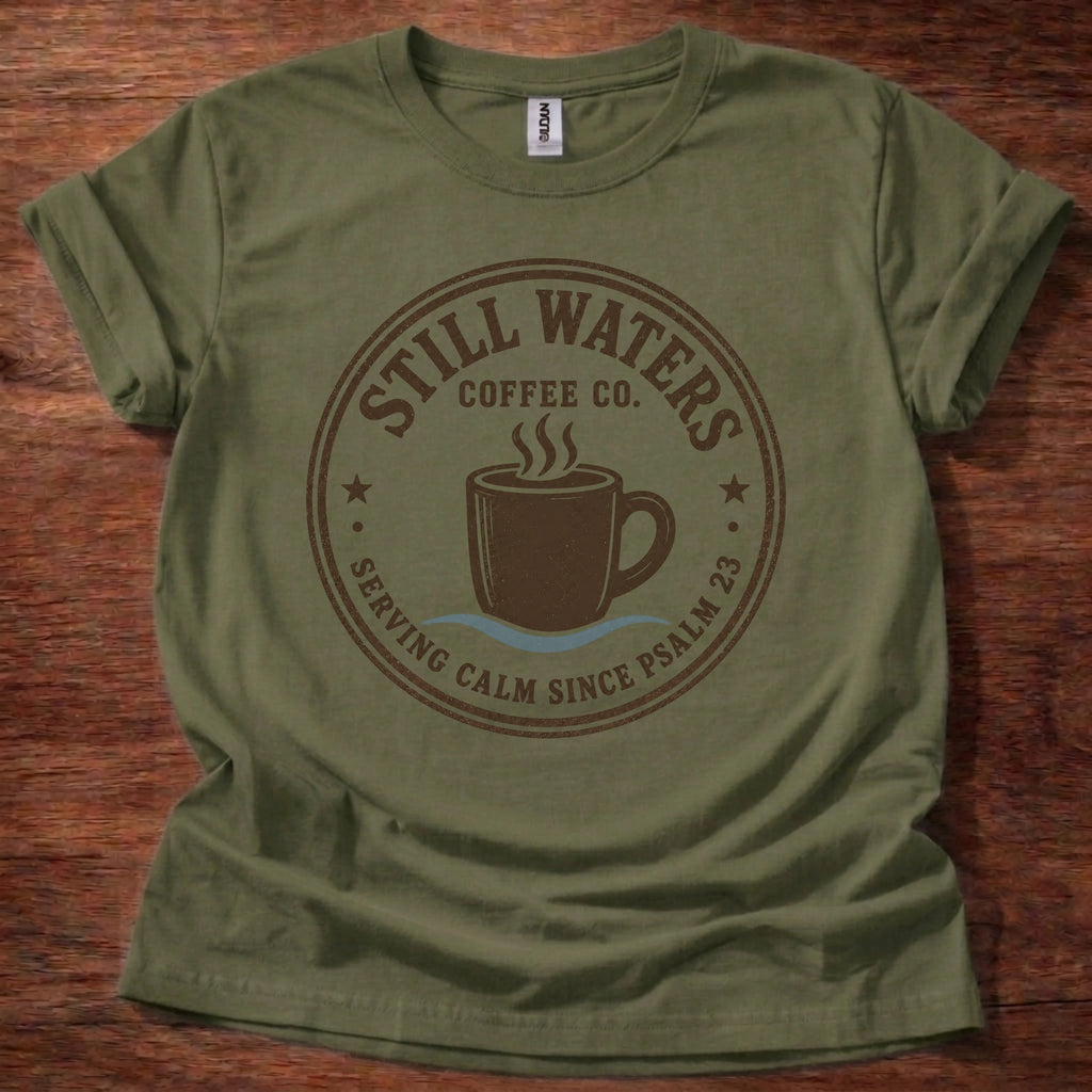 An olive green short-sleeve t-shirt laid flat on a dark wood background, featuring a brown circular graphic that reads 'Still Waters Coffee Co.' with a coffee cup illustration and the phrase 'Serving Calm Since Psalm 23'.