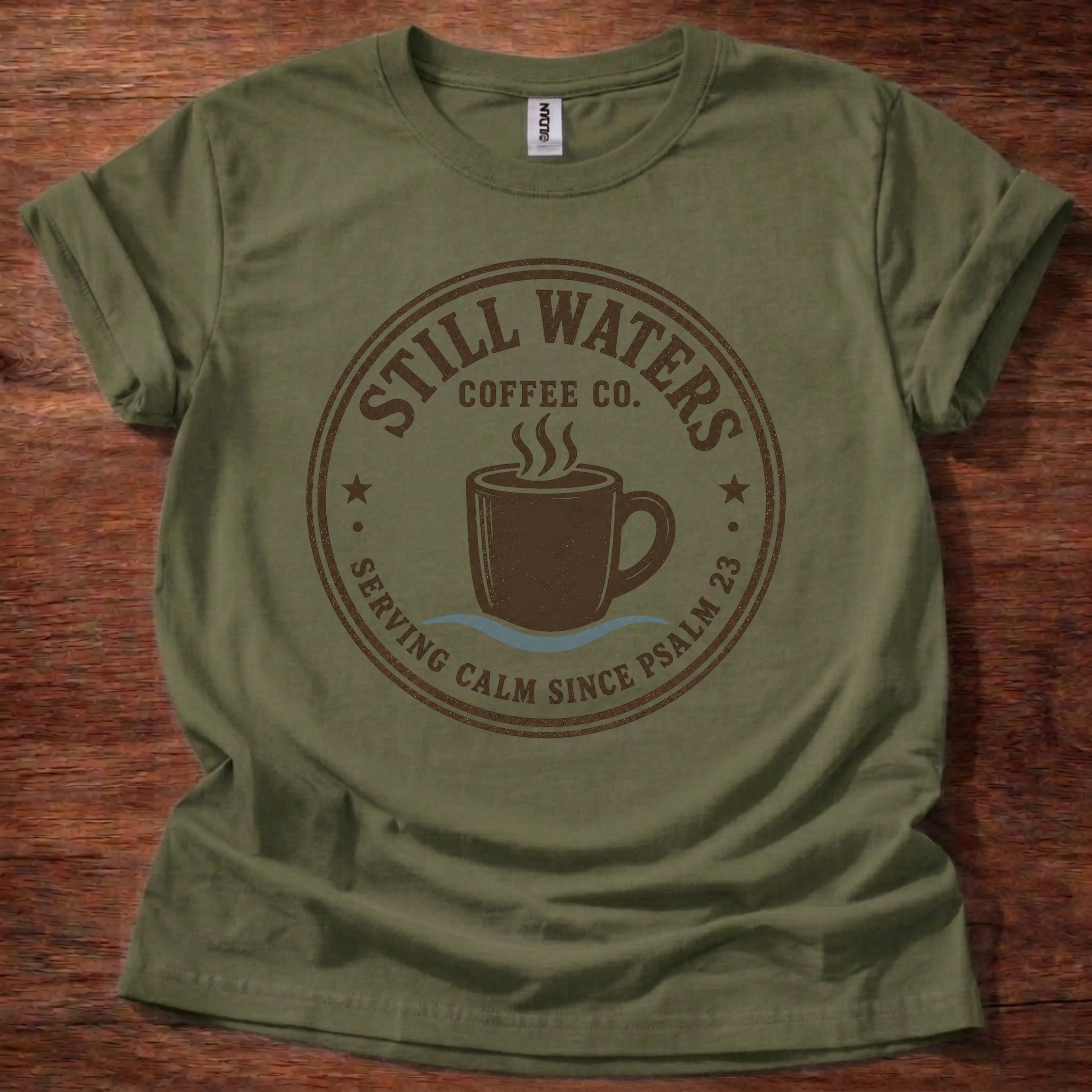 An olive green short-sleeve t-shirt laid flat on a dark wood background, featuring a brown circular graphic that reads 'Still Waters Coffee Co.' with a coffee cup illustration and the phrase 'Serving Calm Since Psalm 23'.