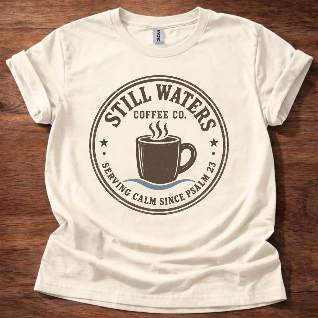 An off-white short-sleeve t-shirt laid flat on a dark wood background, featuring a circular brown graphic print for 'Still Waters Coffee Co.' with a coffee cup illustration and the text 'Serving Calm Since Psalm 23'.