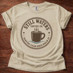A beige short-sleeve t-shirt with a dark brown circular graphic on the front that reads 'Still Waters Coffee Co. Serving Calm Since Psalm 23' around an illustration of a coffee cup, laid flat on a dark wood background.
