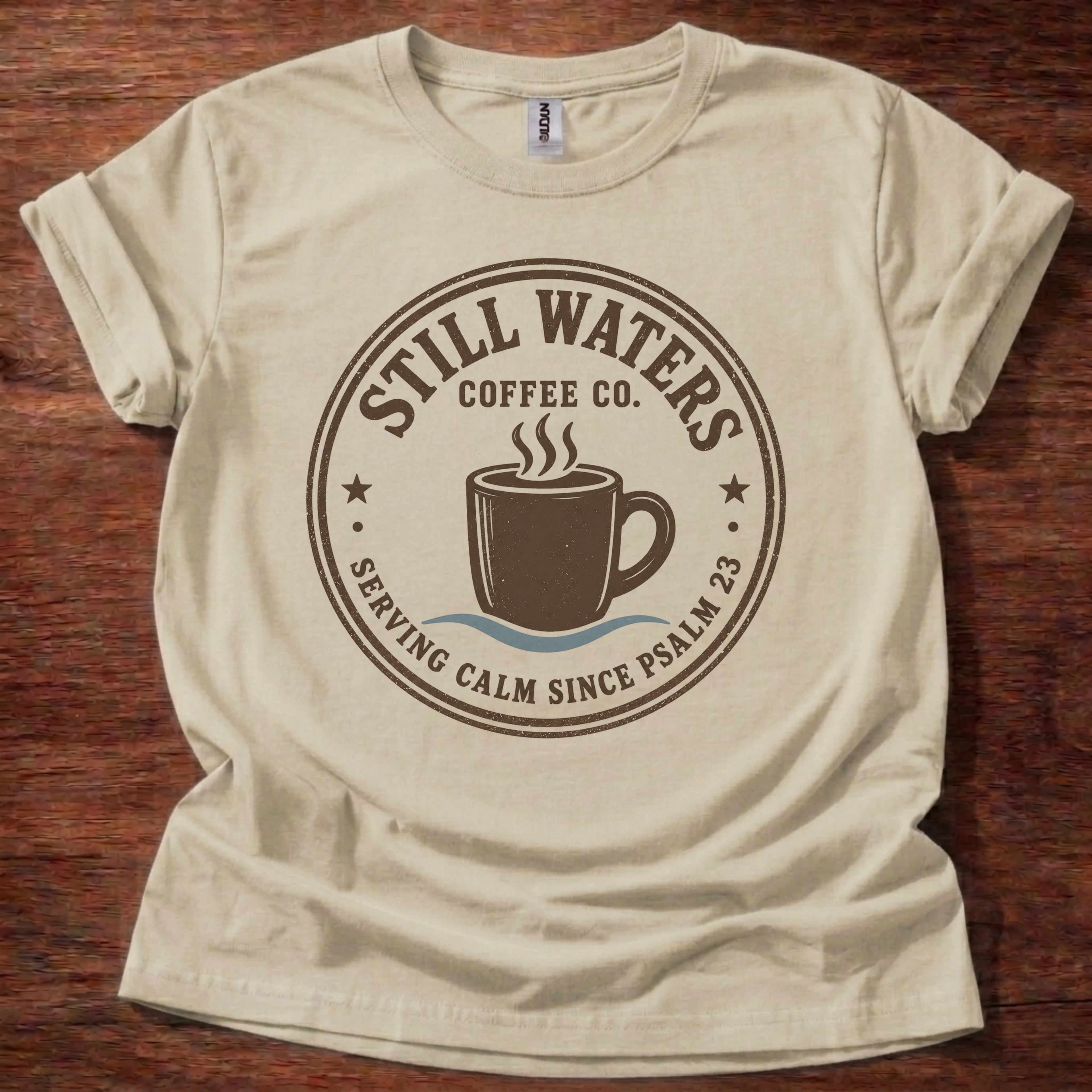 A beige short-sleeve t-shirt with a dark brown circular graphic on the front that reads 'Still Waters Coffee Co. Serving Calm Since Psalm 23' around an illustration of a coffee cup, laid flat on a dark wood background.