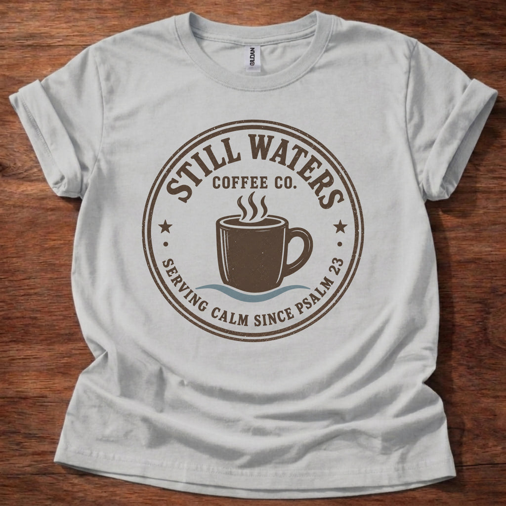 A light gray crewneck t-shirt with a brown circular graphic reading 'Still Waters Coffee Co.' and featuring a coffee cup illustration, laid flat on a dark wood background.