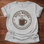 A light gray crewneck t-shirt with a brown circular graphic reading 'Still Waters Coffee Co.' and featuring a coffee cup illustration, laid flat on a dark wood background.