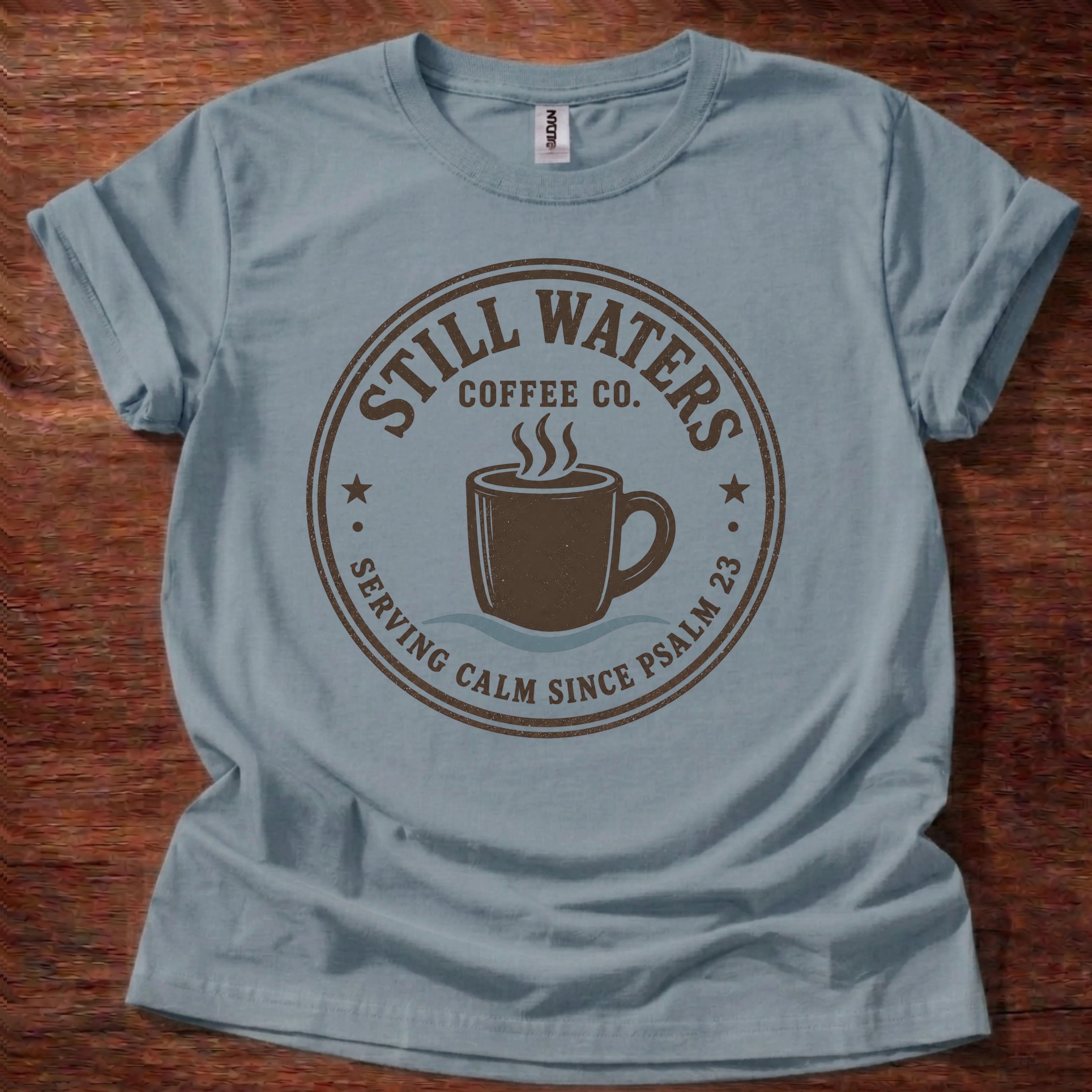 A light blue crewneck t-shirt laid flat on a dark wood background, featuring a brown circular graphic that reads 'Still Waters Coffee Co.' with a coffee cup illustration.