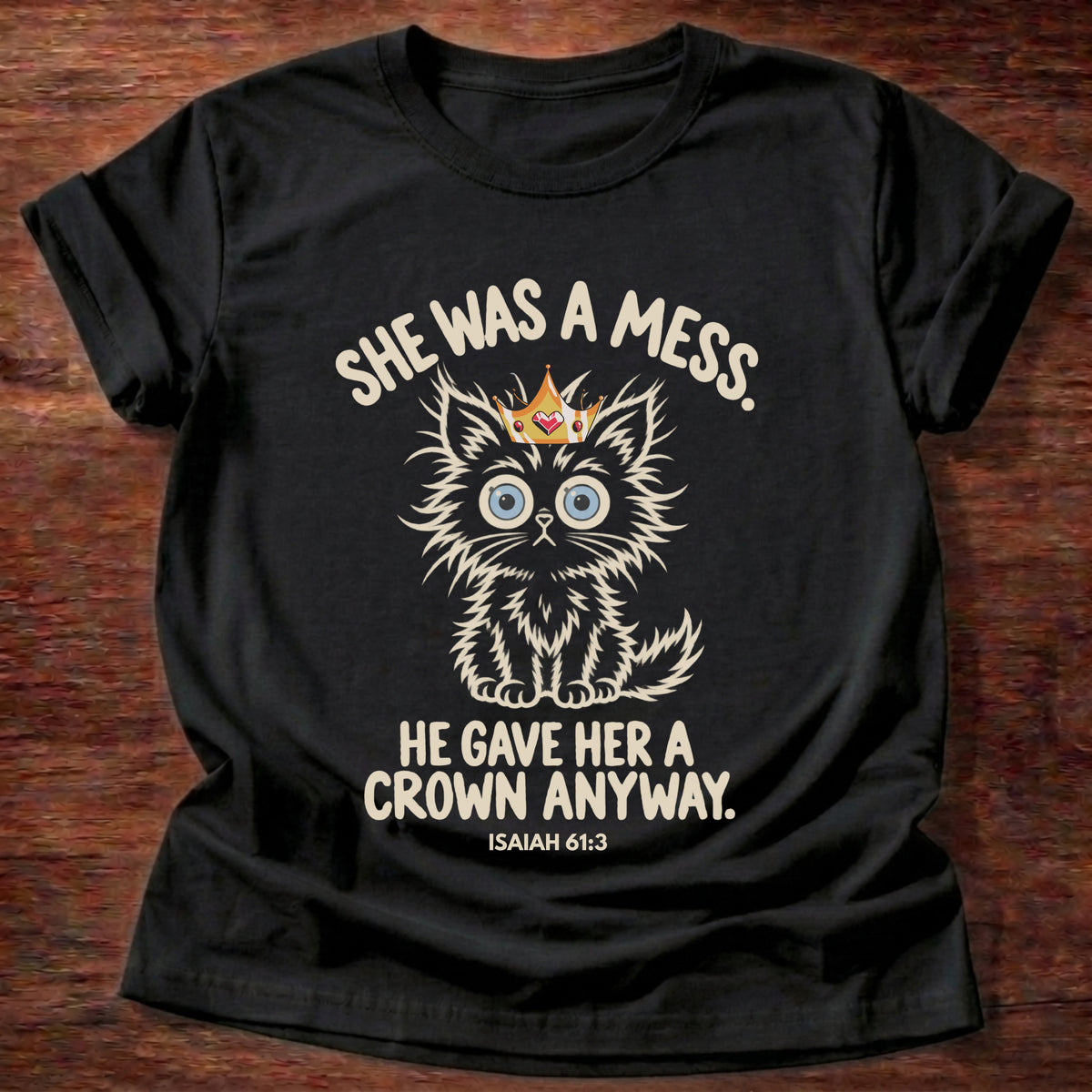 Black t-shirt printed with a fluffy wide-eyed kitten wearing a gold crown graphic and “She was a mess. He gave her a crown anyway.” Isaiah 61:3 Christian shirt