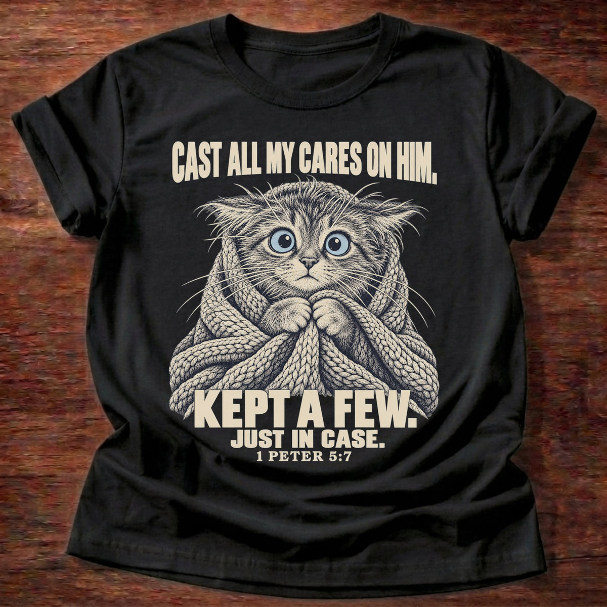 Kept A Few T-Shirt in Black featuring a wide-eyed cat wrapped in a knit blanket and 1 Peter 5:7 anxiety humor design
