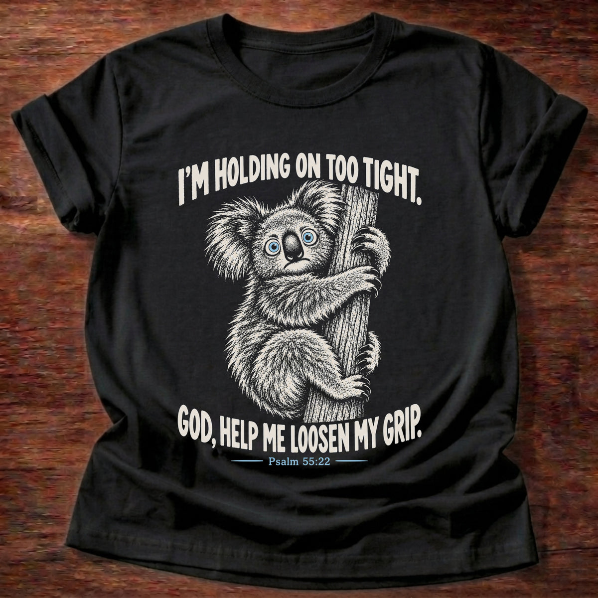 Black t-shirt printed with a wide-eyed koala clinging to a tree trunk graphic and “I'm holding on too tight. God, help me loosen my grip.” Psalm 55:22 faith based gift